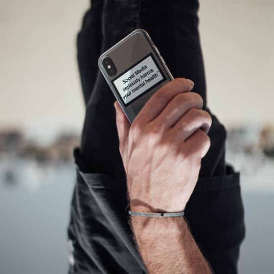 Person is holding their phone upside down. The photo has been flipped round to show the phone the right way up. The person wears black trousers and a black t-shirt. You don't see the whole of their body but just part of their arm and hand holding the phone. The background is blurred our but it looks like they are on a beach with people in the background. We see the phone from the back with the camera showing. It is a sliver phone with a clear protective case. on the case there are words with a black border and white background which say: "Social Media seriously harms your mental health".