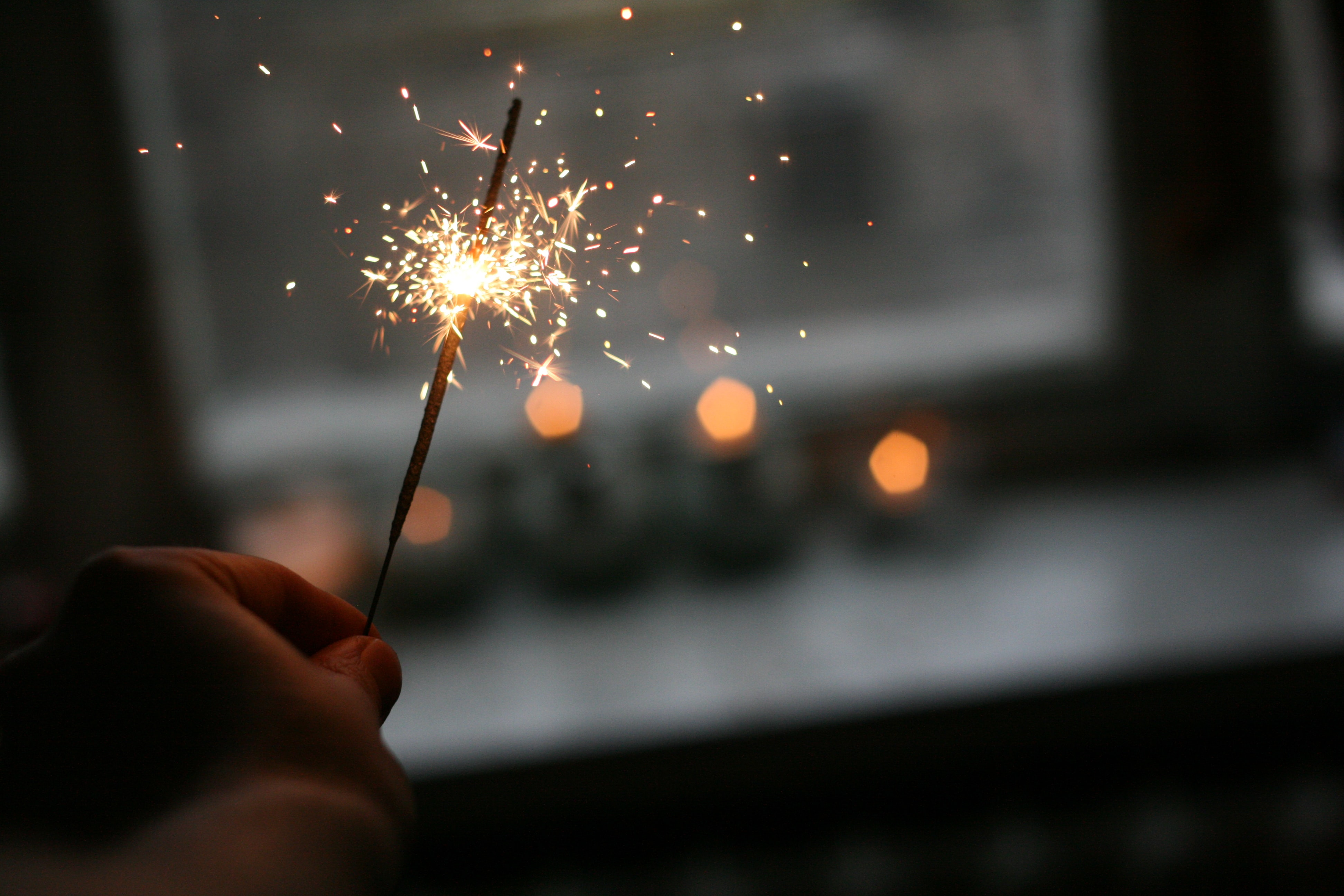 Hand holding a sparkler which is half way through burning. There are sparks flying off the sparkler. The background is blurred but it appears to be that the sparkler is being burned indoors as there is an outline of a window and a windowsill